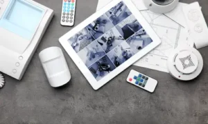Tablet displaying multiple CCTV camera feeds surrounded by security sensors, alarm devices, and control equipment.